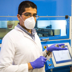 A scientist wearing a lab coat, mask, and gloves operating a laboratory instrument with a blue screen.