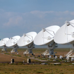 A row of large white satellite dishes under a partly cloudy sky in an open field.