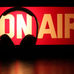 A pair of headphones hangs in front of a glowing red and white "ON AIR" sign in a radio studio.