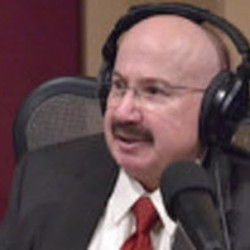 A man with glasses and a mustache wearing headphones and speaking into a microphone in a recording studio.