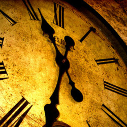 Close-up of an antique clock face showing the time at 11:55 with Roman numerals and a warm, golden glow.