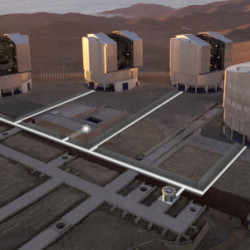 Aerial view of a desert observatory complex with large telescopes mounted on platforms, set against mountainous terrain.