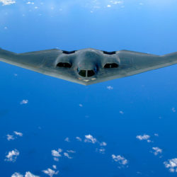 A B-2 Spirit stealth bomber flying over a blue sky with scattered clouds below.