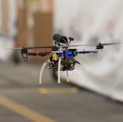 A close-up of a small quadcopter drone hovering indoors with visible wiring and components.