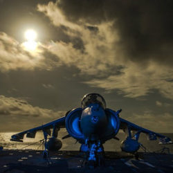 A blue fighter jet on a carrier deck under a dramatic cloudy sky with the sun partially visible.