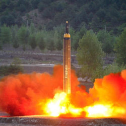 A missile launching from the ground with bright flames and smoke at the base, surrounded by trees and hills.