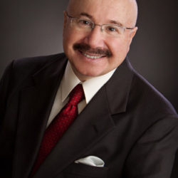 A smiling bald man with glasses wearing a dark suit, white shirt, and red tie against a dark background.