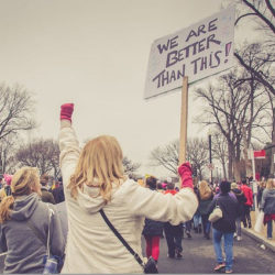 A woman holding a sign that says 'We are better than this!' at a protest or rally with a crowd in the background.
