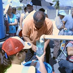A man in an orange shirt signing an autograph for a child wearing a red cap at an outdoor event with people and umbrellas in the background.