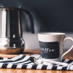 A coffee cup with a chalkboard label reading 'coffee,' placed on a striped cloth next to a spoon and a metallic coffee pot.